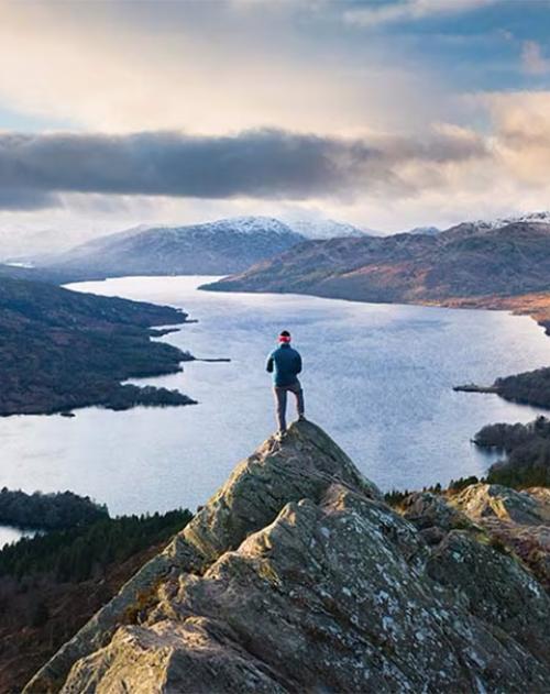 Image of a man standing at the peak of a mountain looking out over a lake and mountains in the distance