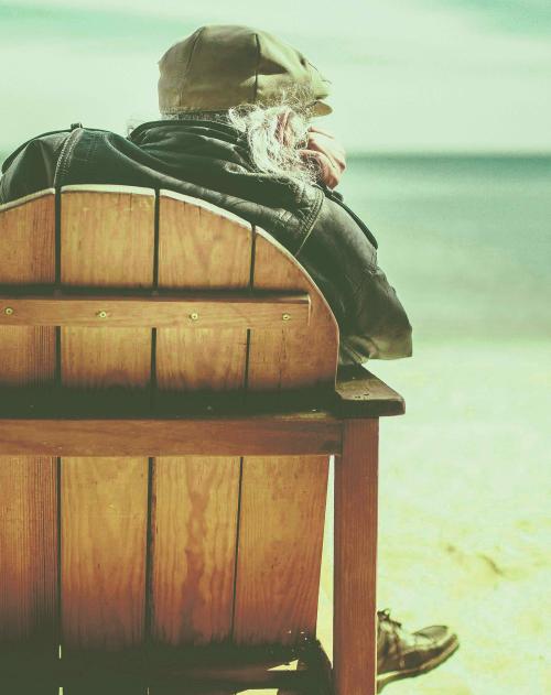 From behind we see an older man with flat cap and long white beard sits in a wooden beach chair in the sand looking out to sea